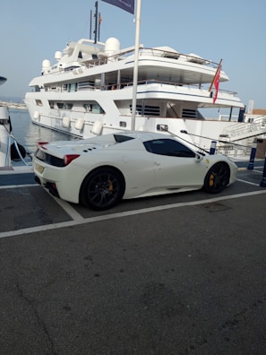 A luxury sports car parked near a large yacht, with both the car and yacht displaying sleek, modern designs. The setting appears to be a marina with calm water and other boats visible in the background.