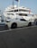 A luxury sports car parked near a large yacht, with both the car and yacht displaying sleek, modern designs. The setting appears to be a marina with calm water and other boats visible in the background.