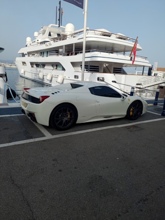 A luxury sports car parked near a large yacht, with both the car and yacht displaying sleek, modern designs. The setting appears to be a marina with calm water and other boats visible in the background.