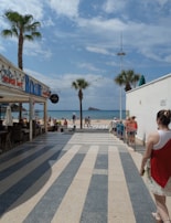 A sunny beach scene features a pathway leading to a sandy shore. There are palm trees, a beach bar with people sitting inside, and various people walking towards the beach. Blue skies and a few clouds are visible above the clear blue sea. An island is seen in the distance.
