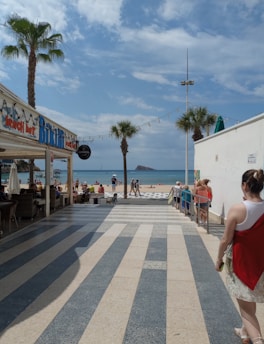 A sunny beach scene features a pathway leading to a sandy shore. There are palm trees, a beach bar with people sitting inside, and various people walking towards the beach. Blue skies and a few clouds are visible above the clear blue sea. An island is seen in the distance.