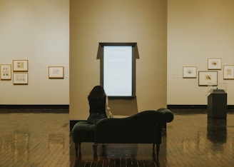 A person sits on a dark green chaise lounge in a gallery or museum, intently viewing a large illuminated information panel on the wall in front of them. Surrounding the panel are framed artworks hung on the light-colored walls. The room has a reflective wooden floor, and the space appears calm with minimalistic decor.