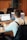 A person wearing a white shirt with black suspenders is seated at a desk, working on two laptop computers. A takeaway coffee cup is placed on the desk, featuring a logo. The background includes office chairs and a wooden panel wall with plants visible.