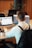 A person wearing a white shirt with black suspenders is seated at a desk, working on two laptop computers. A takeaway coffee cup is placed on the desk, featuring a logo. The background includes office chairs and a wooden panel wall with plants visible.
