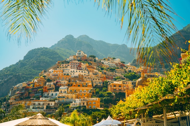 A picturesque hillside town featuring colorful stacked buildings with a background of lush green mountains. Palm leaves frame the top of the image, and an umbrella is visible in the foreground, suggesting a tropical atmosphere.