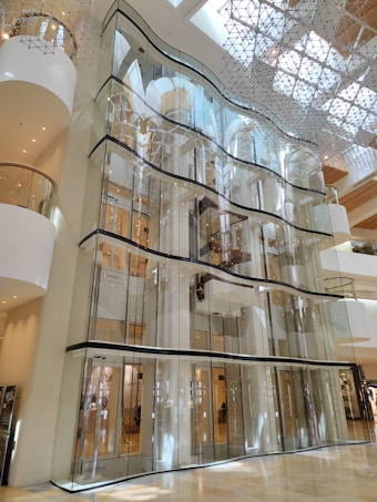 A multi-story structure with a modern architectural design featuring large glass panels and a spacious, open interior. The upper levels showcase artistic, geometric installations on the ceiling, allowing natural light to flood through. The building has a luxurious and sleek aesthetic, dominated by reflective surfaces and soft lighting.