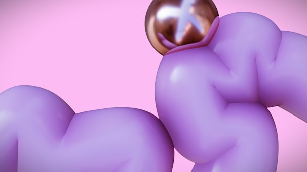 A surreal image featuring two purple, inflated, abstract shapes against a pink background. A shiny, metallic, spherical object with reflective properties is situated at the top of one of the shapes.