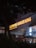 A dimly lit storefront featuring illuminated signs for Popeyes and Burger King. The signage is prominent with bright orange lettering against a dark background. The scene includes umbrellas and an entrance area, suggesting a shared location or food court setting.