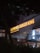 A dimly lit storefront featuring illuminated signs for Popeyes and Burger King. The signage is prominent with bright orange lettering against a dark background. The scene includes umbrellas and an entrance area, suggesting a shared location or food court setting.