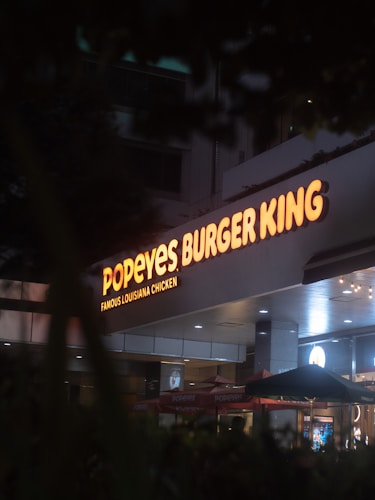 A dimly lit storefront featuring illuminated signs for Popeyes and Burger King. The signage is prominent with bright orange lettering against a dark background. The scene includes umbrellas and an entrance area, suggesting a shared location or food court setting.