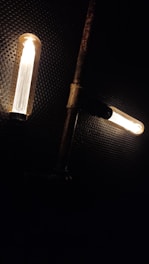 Two vintage-style light bulbs are illuminated against a dark, perforated background. The bulbs are horizontally aligned and connected to a rusty metal pipe structure, creating a steampunk aesthetic.