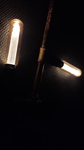 Two vintage-style light bulbs are illuminated against a dark, perforated background. The bulbs are horizontally aligned and connected to a rusty metal pipe structure, creating a steampunk aesthetic.