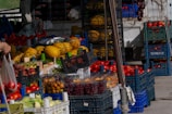 A market stall displaying a variety of fresh fruits and vegetables, including melons, tomatoes, cherries, grapes, and other produce. Fruits are organized in crates and baskets, with vibrant colors suggesting freshness. A hand is holding a plastic bag containing tomatoes.