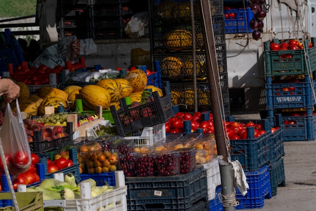 A market stall displaying a variety of fresh fruits and vegetables, including melons, tomatoes, cherries, grapes, and other produce. Fruits are organized in crates and baskets, with vibrant colors suggesting freshness. A hand is holding a plastic bag containing tomatoes.