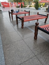 Several traditional woven beds, known as charpoys, are arranged on a tiled outdoor area. A person is lying on one of the beds, looking relaxed, with a backdrop of red and white signage. Small, well-manicured bushes and trees are visible in the background.
