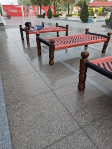 Several traditional woven beds, known as charpoys, are arranged on a tiled outdoor area. A person is lying on one of the beds, looking relaxed, with a backdrop of red and white signage. Small, well-manicured bushes and trees are visible in the background.