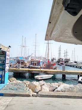 A marina filled with various boats and yachts docked at a pier. The scene includes masts, flags, and a small motorboat in the foreground. The water is a clear blue, and there are rocks lining the shore next to the road. A kiosk or booth advertising daily boat trips is visible on the left.