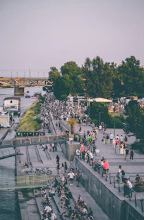 A bustling riverside area filled with people walking, sitting, and socializing. The scene features a mix of urban and natural elements, with trees lining the pathway and a bridge crossing the river in the background. Bicycles are seen parked along the pathway, adding to the urban vibe of the setting.
