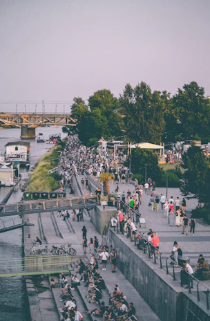 A bustling riverside area filled with people walking, sitting, and socializing. The scene features a mix of urban and natural elements, with trees lining the pathway and a bridge crossing the river in the background. Bicycles are seen parked along the pathway, adding to the urban vibe of the setting.