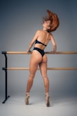 A dancer in a black leotard and pointe shoes stands at a ballet barre with her back arched and head tilted back, causing her hair to flow dramatically. The setting is minimalistic, with a neutral gray background.