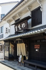 A traditional Japanese storefront featuring wooden architectural details and a prominent sign with Japanese characters. The building has sliding doors with fabric curtains partially covering the entrance, typical of traditional shops. A bench and a small sign with images are placed by the entrance.
