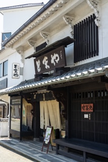 A traditional Japanese storefront featuring wooden architectural details and a prominent sign with Japanese characters. The building has sliding doors with fabric curtains partially covering the entrance, typical of traditional shops. A bench and a small sign with images are placed by the entrance.