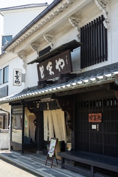 A traditional Japanese storefront featuring wooden architectural details and a prominent sign with Japanese characters. The building has sliding doors with fabric curtains partially covering the entrance, typical of traditional shops. A bench and a small sign with images are placed by the entrance.