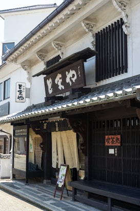 A traditional Japanese storefront featuring wooden architectural details and a prominent sign with Japanese characters. The building has sliding doors with fabric curtains partially covering the entrance, typical of traditional shops. A bench and a small sign with images are placed by the entrance.