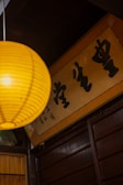 A warm-toned interior with a yellow paper lantern hanging prominently in the foreground. Below the lantern, a wooden panel wall is visible, along with a framed sign featuring bold black calligraphy characters.