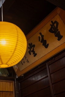 A warm-toned interior with a yellow paper lantern hanging prominently in the foreground. Below the lantern, a wooden panel wall is visible, along with a framed sign featuring bold black calligraphy characters.