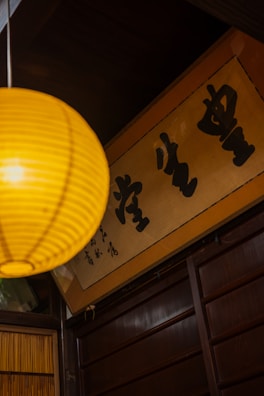 A warm-toned interior with a yellow paper lantern hanging prominently in the foreground. Below the lantern, a wooden panel wall is visible, along with a framed sign featuring bold black calligraphy characters.