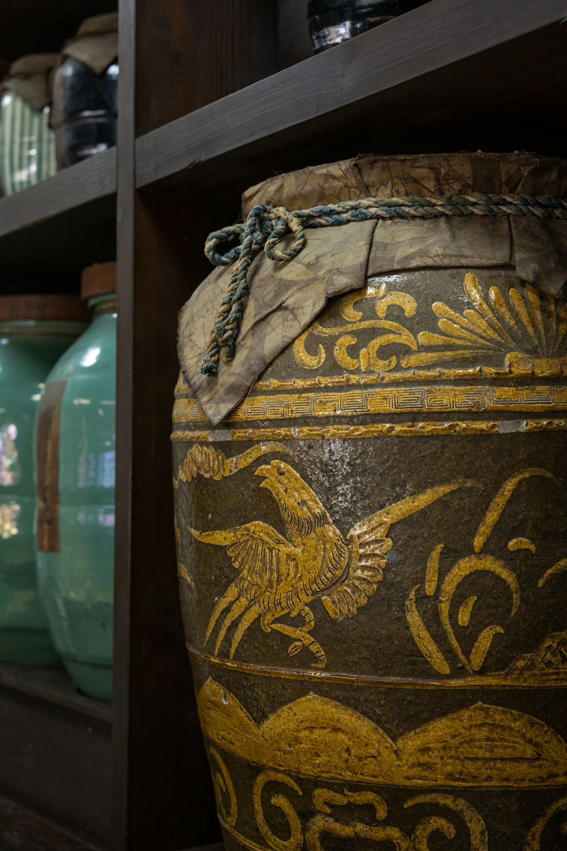 A large, intricately designed ceramic jar features golden ornamental patterns, including a stylized bird, set against a dark, possibly earthenware background. The jar is sealed with a cloth cover tied with a rope. In the background, there are other jars in shades of green and brown, displayed on wooden shelves.