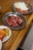 A wooden table with three metal plates containing various uncooked cuts of marbled beef and a side dish of kimchi and pickled vegetables.