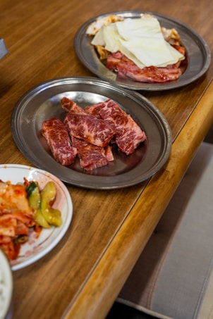 A wooden table with three metal plates containing various uncooked cuts of marbled beef and a side dish of kimchi and pickled vegetables.
