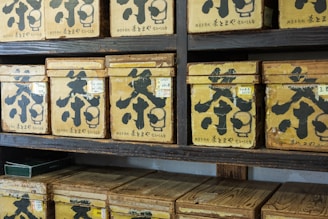 Multiple vintage wooden boxes with Japanese characters are arranged on dark wooden shelves. Each box is labeled with large black calligraphy and smaller labels, giving the appearance of a traditional tea or spice storage area.