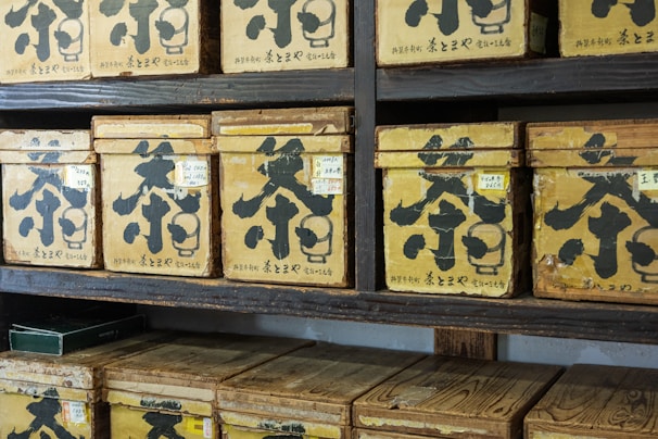 Multiple vintage wooden boxes with Japanese characters are arranged on dark wooden shelves. Each box is labeled with large black calligraphy and smaller labels, giving the appearance of a traditional tea or spice storage area.