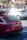 A red Audi with a unique license plate is parked on a street. The car has a roof box attached. The background shows a sunny day with other vehicles and buildings in an urban setting.