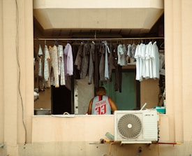 An outdoor scene shows clothes hanging on a line, including white t-shirts and darker clothing, possibly pants. A person in a jersey with the number 33 is visible inside a window. Below the window, an air conditioning unit is mounted on the wall.
