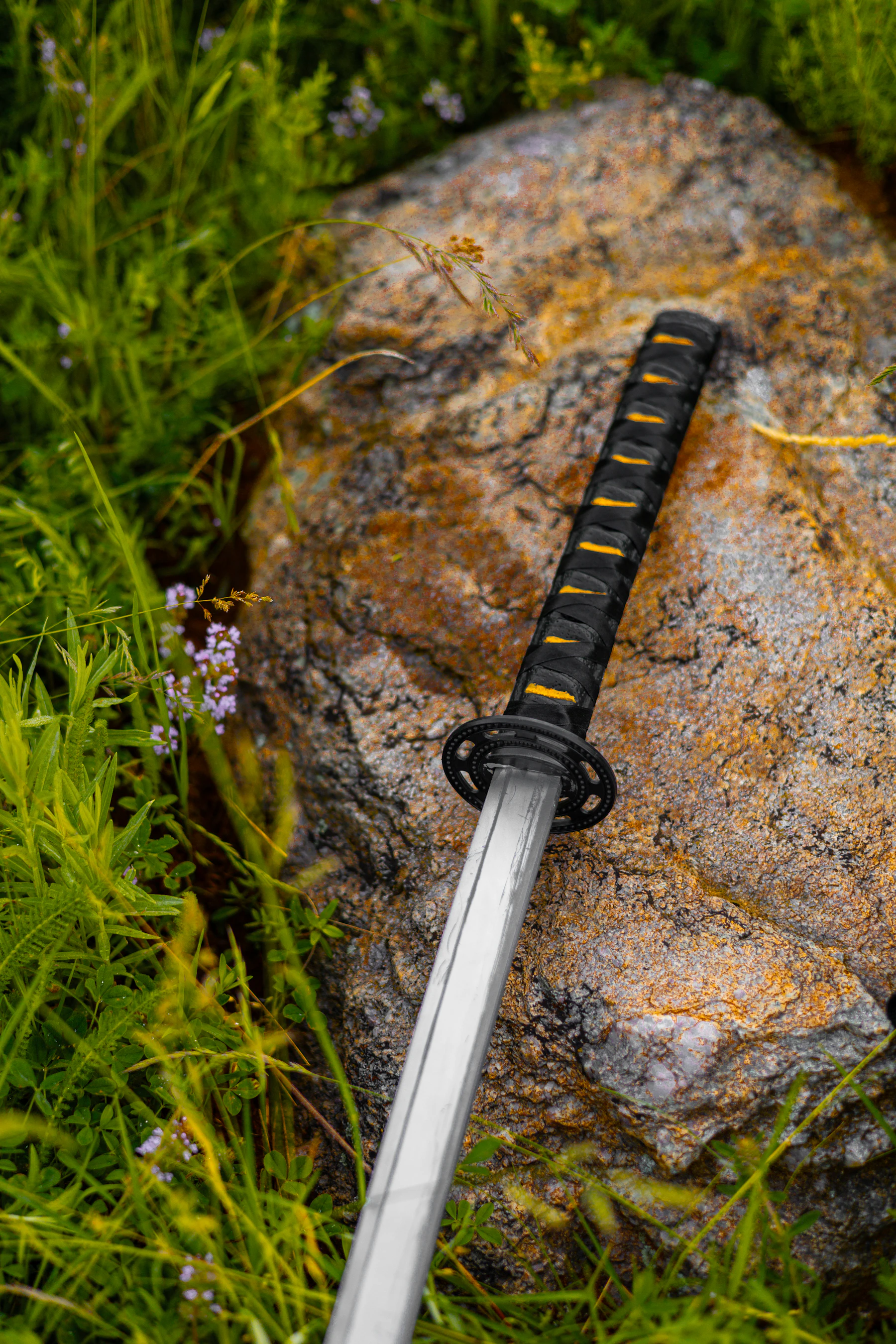 A katana with a black and gold wrapped grip and a round guard lying on a lichen-covered rock