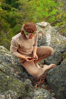 A person with curly hair is wearing a wreath of flowers on their head and dressed in earthy-toned clothing. They are sitting on a rock surrounded by green moss and foliage, examining pine cones in their hands. The environment is a forest with abundant greenery.