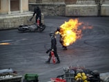A person engulfed in fire walks on a paved ground, accompanied by two other individuals holding fire extinguishers. A motorcycle lies on its side near a stone wall in the background. Various containers and objects are present in the foreground.