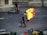 A person engulfed in fire walks on a paved ground, accompanied by two other individuals holding fire extinguishers. A motorcycle lies on its side near a stone wall in the background. Various containers and objects are present in the foreground.