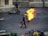 A person engulfed in fire walks on a paved ground, accompanied by two other individuals holding fire extinguishers. A motorcycle lies on its side near a stone wall in the background. Various containers and objects are present in the foreground.