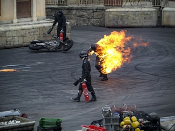 A person engulfed in fire walks on a paved ground, accompanied by two other individuals holding fire extinguishers. A motorcycle lies on its side near a stone wall in the background. Various containers and objects are present in the foreground.