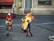A person wearing a fire-resistant suit is engulfed in flames, participating in a controlled stunt or emergency procedure. Surrounding them are individuals in firefighting gear, each holding fire extinguishers and standing ready to intervene. The background includes industrial and possibly urban elements, with a building that has store-like features including a window displaying some items.