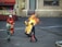 A person wearing a fire-resistant suit is engulfed in flames, participating in a controlled stunt or emergency procedure. Surrounding them are individuals in firefighting gear, each holding fire extinguishers and standing ready to intervene. The background includes industrial and possibly urban elements, with a building that has store-like features including a window displaying some items.