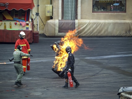 A person wearing a fire-resistant suit is engulfed in flames, participating in a controlled stunt or emergency procedure. Surrounding them are individuals in firefighting gear, each holding fire extinguishers and standing ready to intervene. The background includes industrial and possibly urban elements, with a building that has store-like features including a window displaying some items.