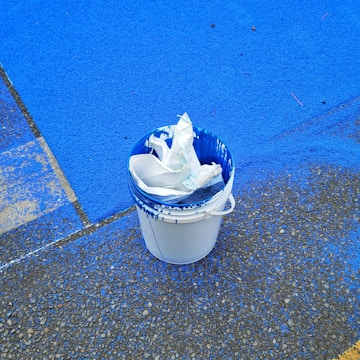 A white bucket partially filled with used plastic paint liners is set on a textured ground. Blue paint has spilled around the bucket on the pavement, creating vivid splashes and stains.