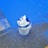 A white bucket partially filled with used plastic paint liners is set on a textured ground. Blue paint has spilled around the bucket on the pavement, creating vivid splashes and stains.