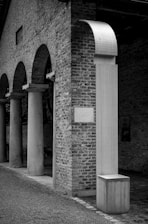 An architectural scene featuring a series of stone columns and arches made of brick. A modern sculpture with a curved top is prominently displayed next to the columns, blending contemporary and historical styles.